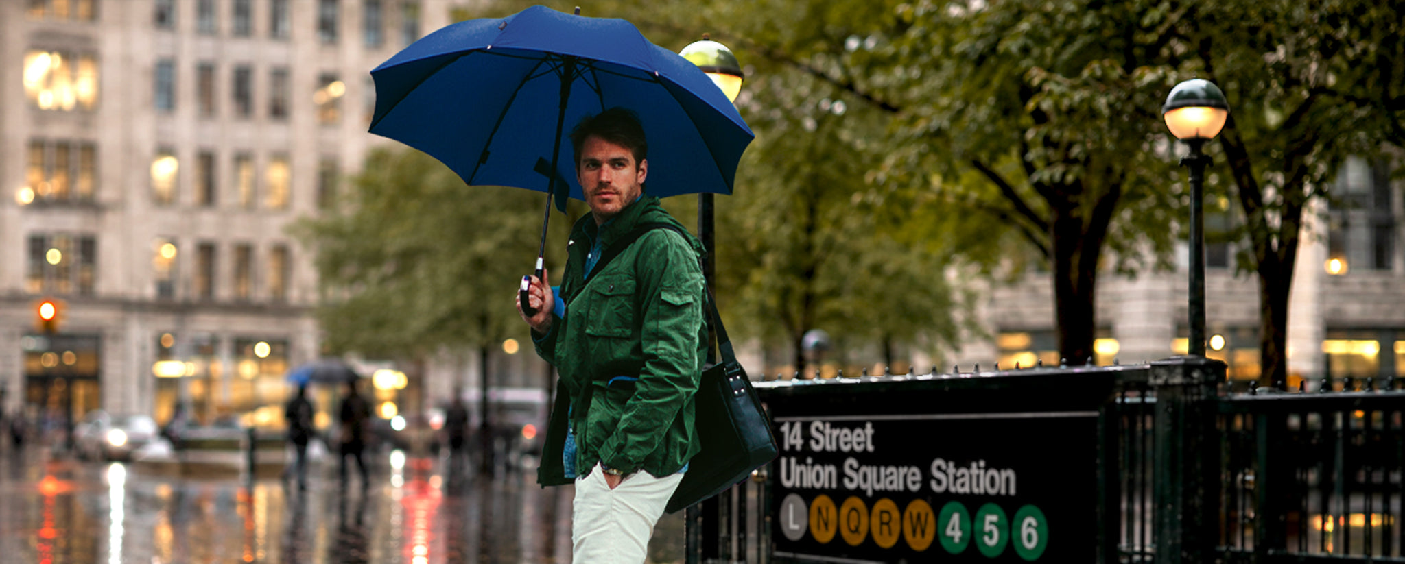 A man standing in the rain at a subway entrance holding a dark blue umbrella, wearing a green jacket, white pants, and carrying a black shoulder bag. The background shows a blurred city street scene with buildings, trees, and a New York City subway sign.