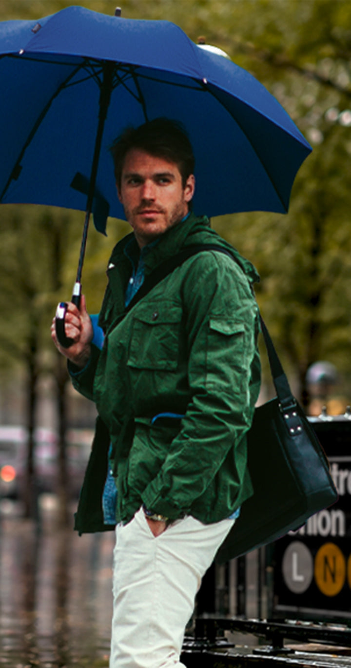 A man standing in the rain at a subway entrance holding a dark blue umbrella, wearing a green jacket, white pants, and carrying a black shoulder bag. The background shows a blurred city street scene with buildings, trees, and a New York City subway sign.