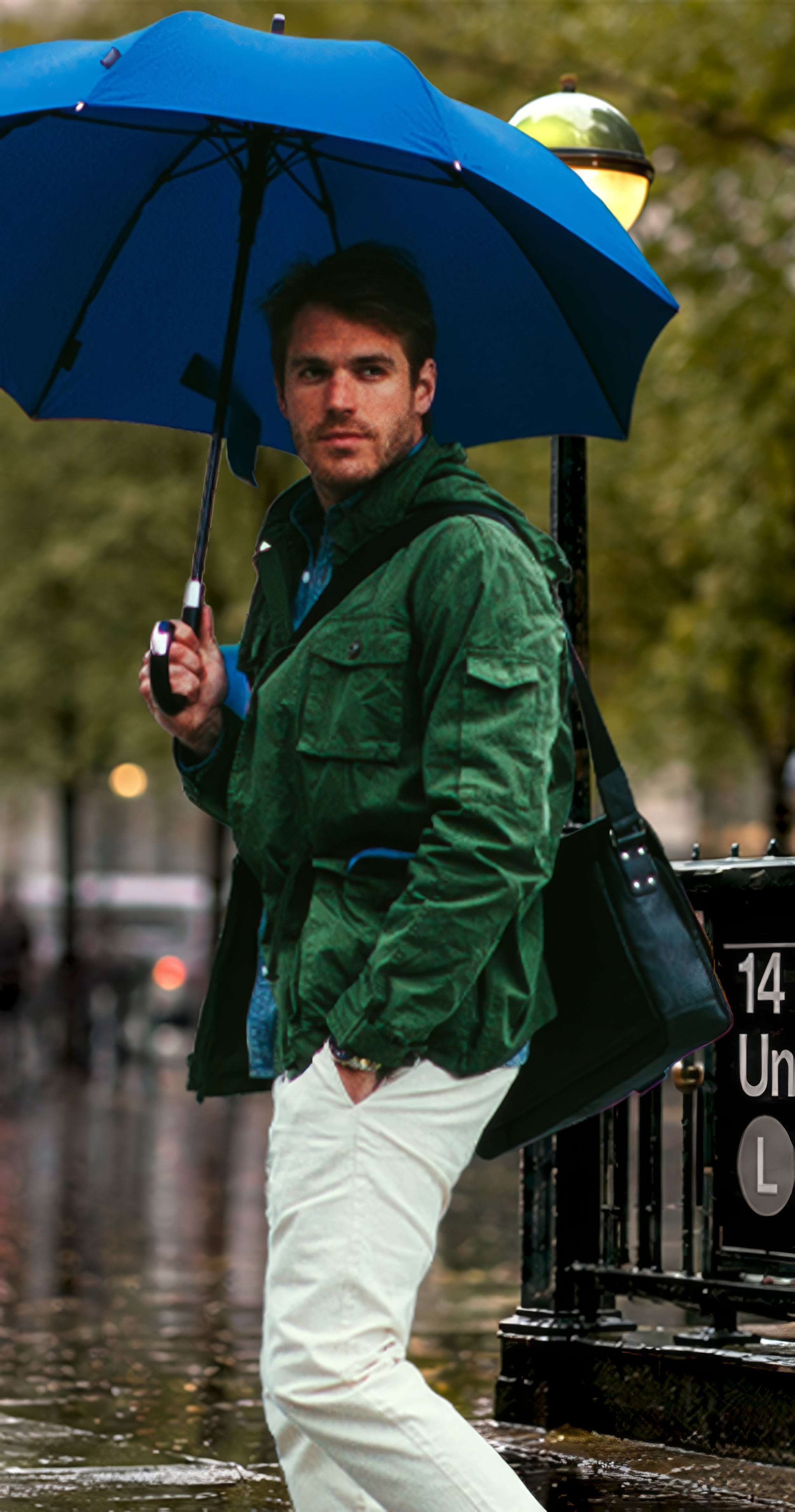 A man standing outdoors in the rain holding a large blue umbrella, wearing a green jacket, white pants, and carrying a dark shoulder bag.